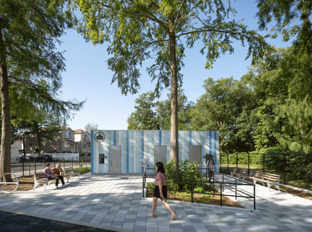 Wide-angle view of the Staten Island public restroom at Lopez Playground, showcasing its blue-striped tile facade, accessible ramp, and surrounding park seating under leafy trees on a sunny day.