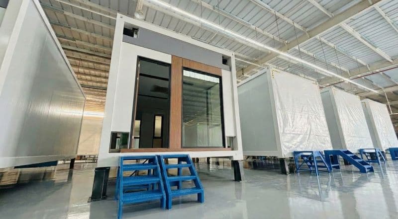 Factory interior showing prefabricated modular units for the Yaound&eacute; office building, with one unit displayed on blue supports under industrial lighting.
