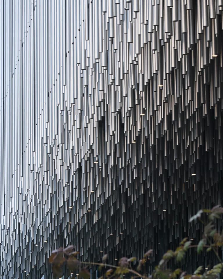 Close-up of textured aluminum pipe facade with integrated linear lighting enhancing material rhythm at dusk.
