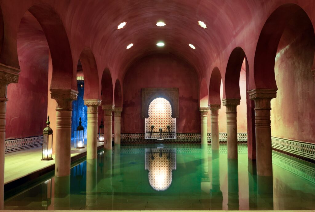Interior of a restored historic hammam in Marrakech or Fez, featuring vaulted terracotta ceilings, arched colonnades, and a reflective pool with ornate tilework  demonstrating Islamic architecture&rsquo;s integration of thermal comfort, light modulation, and spiritual ambiance.