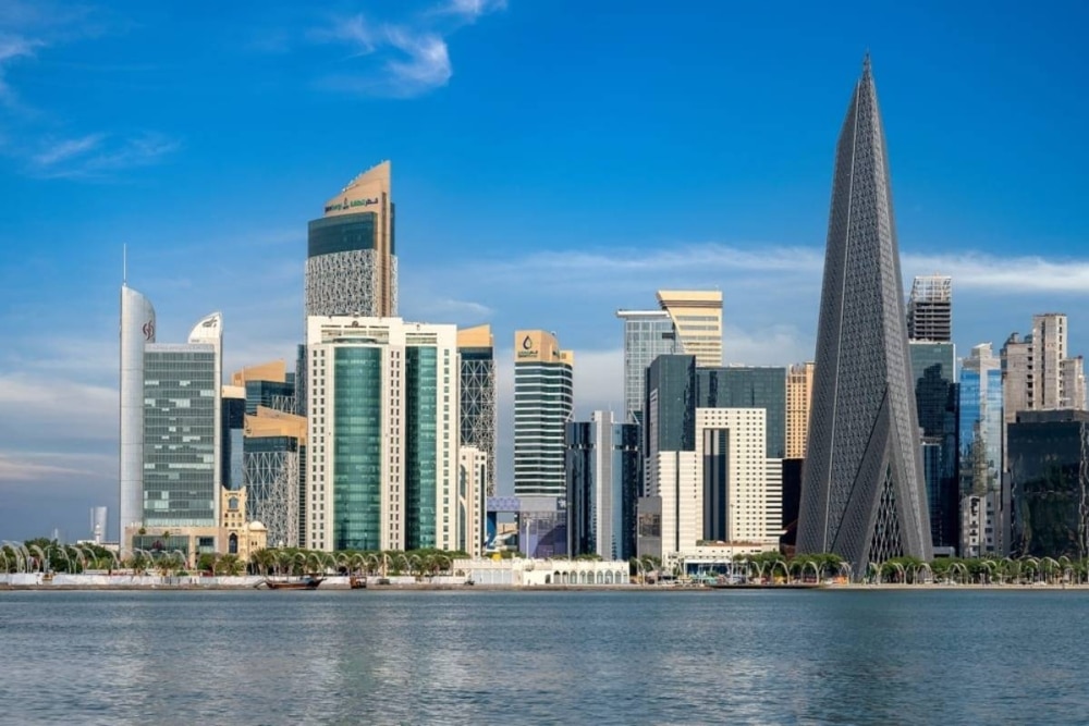 A panoramic view of the modern Doha skyline in Qatar featuring unique skyscrapers like the Burj Doha and the Doha Tower under a clear blue sky, viewed from across the water.