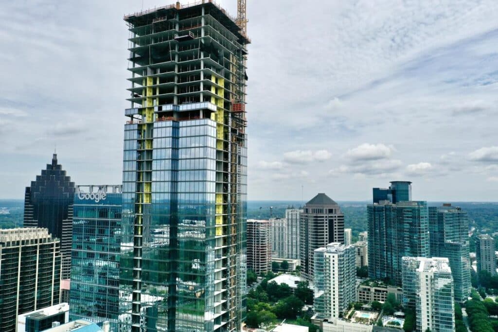 Construction crane beside partially clad high-rise, reflecting historic building in its glass facade under overcast sky.