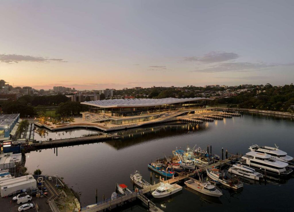 Evening aerial view  at Blackwattle Bay, illuminated against twilight skies, with fishing boats docked nearby and urban skyline in background.