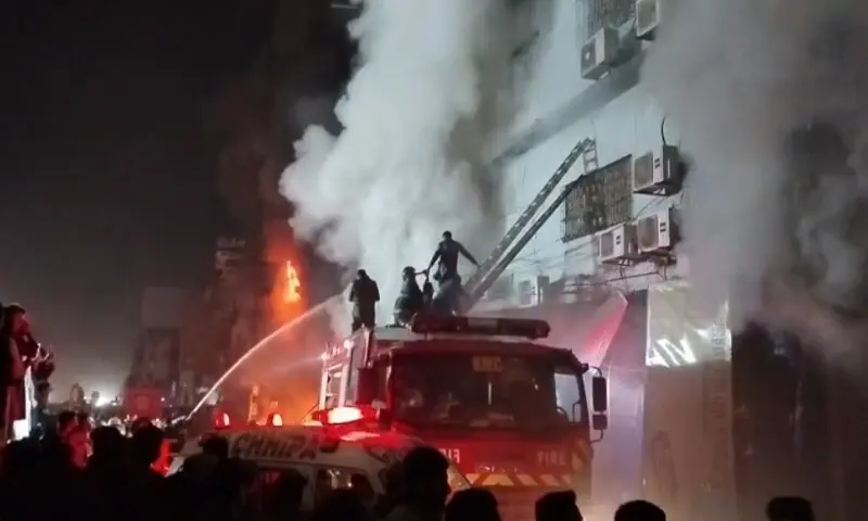  Firefighters on a ladder truck battle flames erupting from upper floors of a commercial building during the January 18, 2026 Karachi shopping center fire, as thick smoke billows into the night sky amid crowd gathering below.