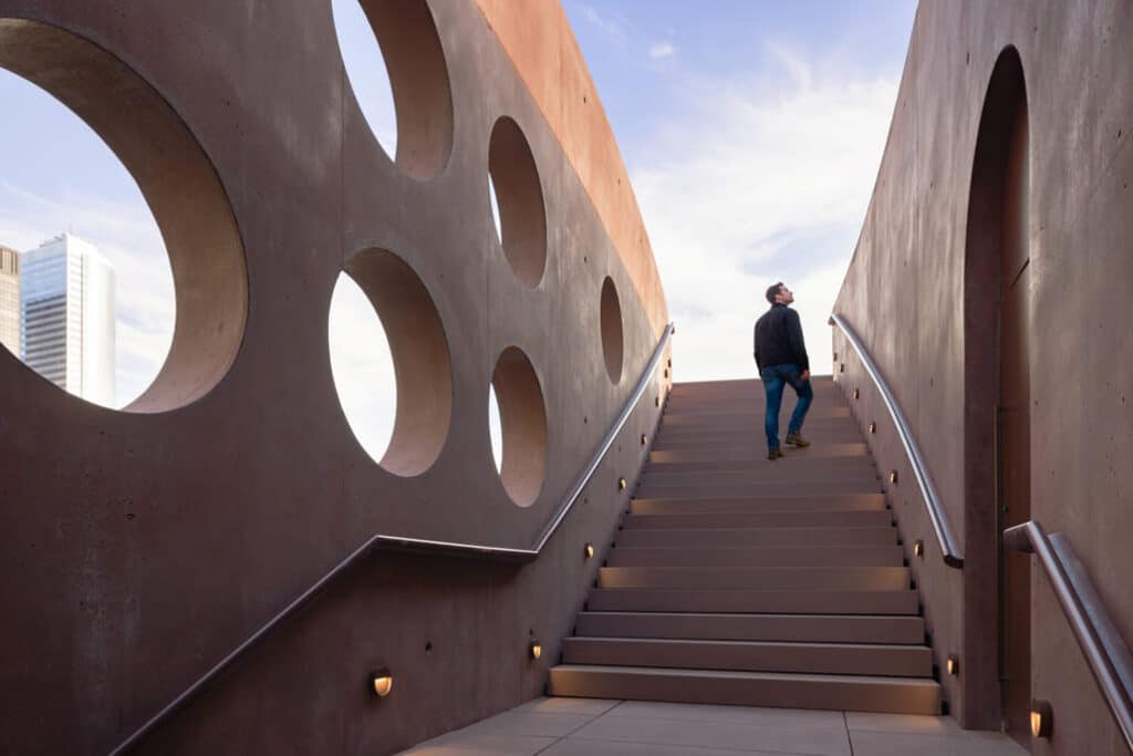A visitor ascends the wide, rust-toned concrete staircase of Wagner Park Pavilion in Lower Manhattan, framed by circular apertures and minimalist railings under a clear sky &mdash; highlighting its sculptural form and public accessibility.