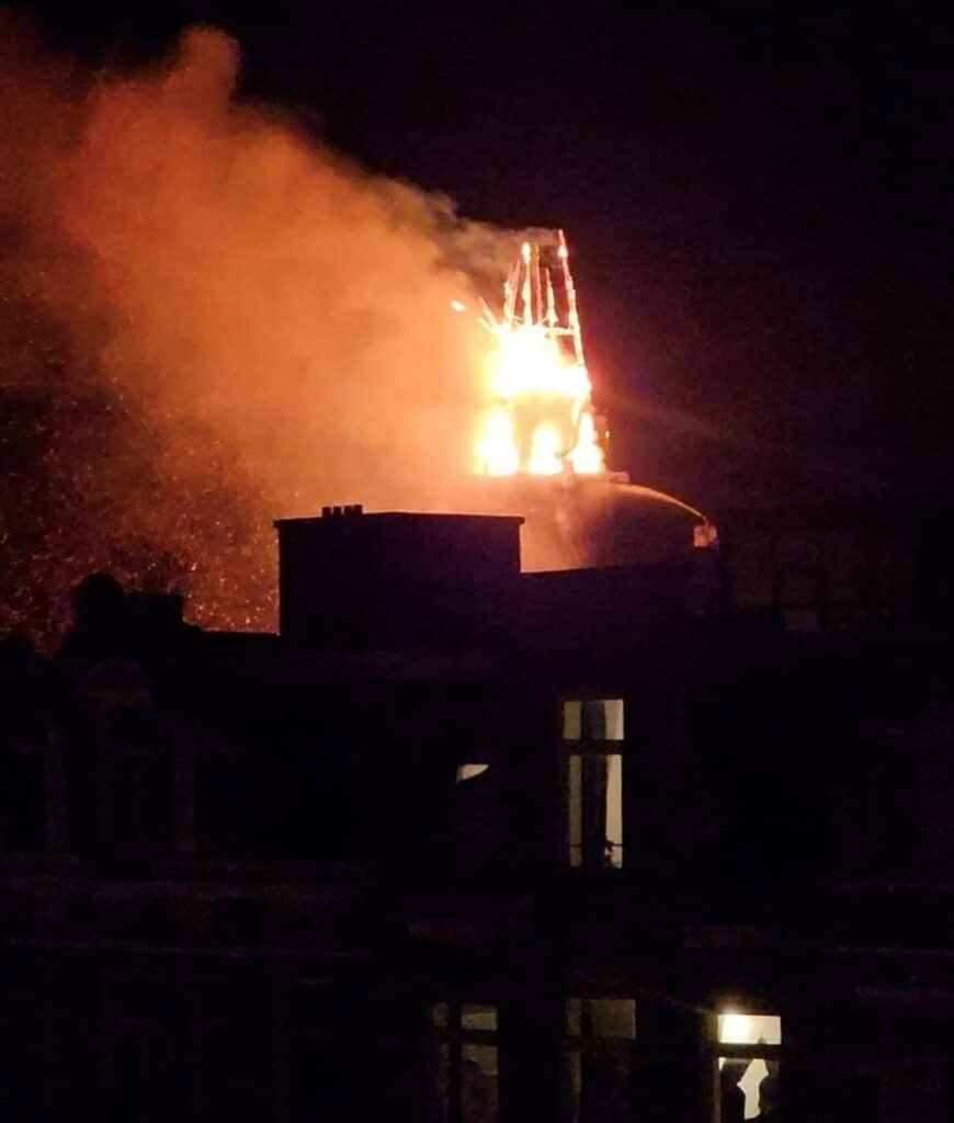 Flames erupt from the roof of a historic building at night, illuminating smoke against the dark sky as surrounding structures remain in shadow.