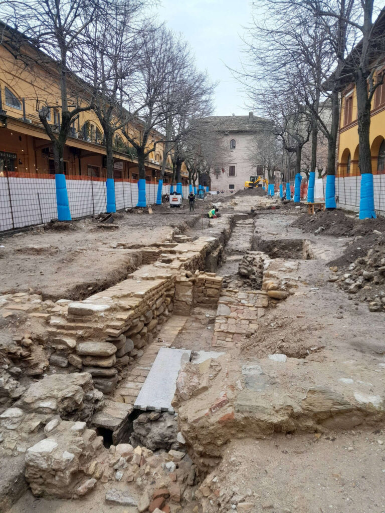 A wide view of the archaeological excavation site in Fano, showing the extensive foundations of the Roman basilica running along a modern street