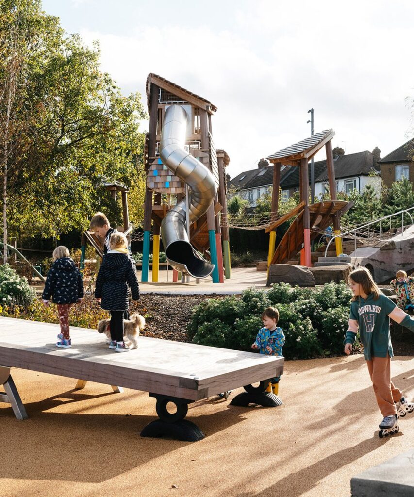 A wide view of children enjoying the various wooden play structures and the large metal slide at the newly designed park in Southwark.