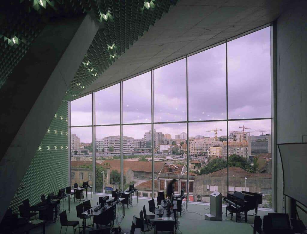 منظر من إحدى المساحات العامة داخل كازا دا موزيكا، يُظهر طاولات وكراسي خلف واجهة زجاجية مموجة وكبيرة تطل على المدينة.