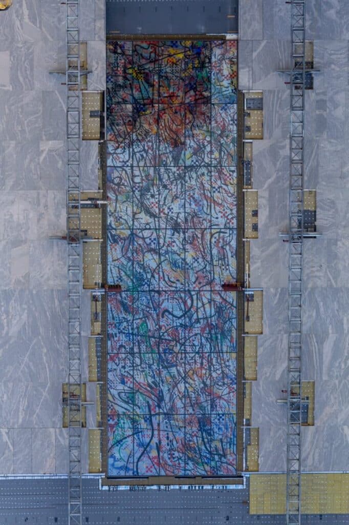 منظر جوي لجدارية مجردة كبيرة مثبتة على سطح مبنى قيد الإنشاء، محاطة بسقالات وأطر هيكلية.