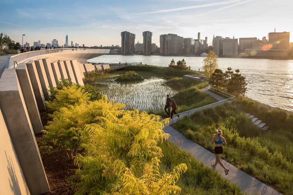 A landscaped waterfront park with winding paths and native vegetation, set against the Manhattan skyline at sunset, demonstrating adaptive reuse of urban edge space.