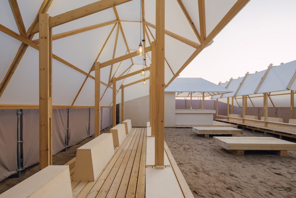 Close up of exposed timber framing and pendant bulbs inside a temporary pavilion, showing structural logic and material honesty.