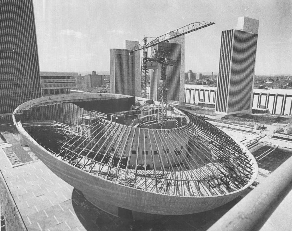 Steel framework of a large dome-shaped building under construction, surrounded by mid-century high-rise towers.