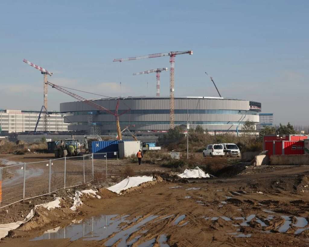  Exterior view of the near-completed Milan 2026 Olympic Ice Hockey Arena in Santa Giulia, with cranes still active and surrounding construction site under a clear sky.