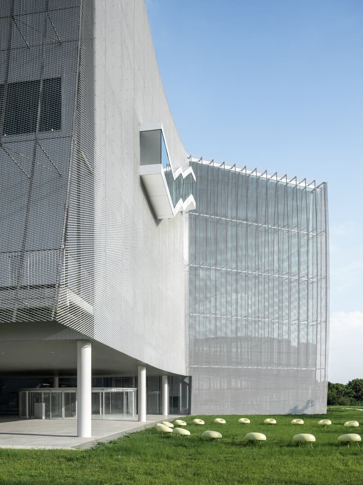 Close-up of the building&rsquo;s textured facade with expanded metal mesh and angular glass protrusions, set against a clear sky and landscaped lawn.