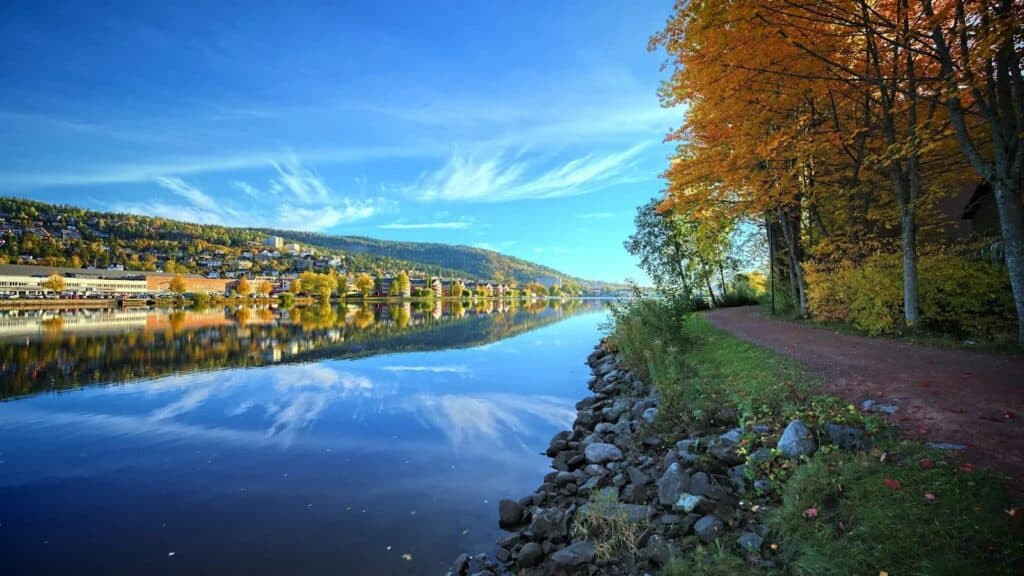 Drammen River Park&rsquo;s autumn pathway along the riverbank, with tree reflections on calm water and hillside urban fabric in background.