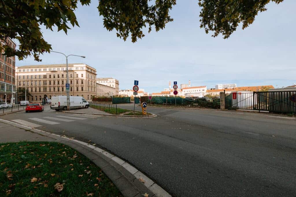 Street view of the future site for the New Brno Municipal Building, showing existing civic buildings, road signage, and perimeter fencing under a clear sky.