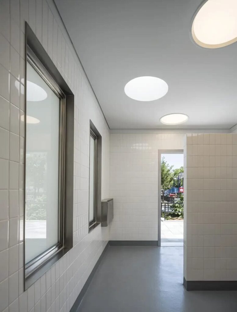 Interior view of the Staten Island public restroom at Lopez Playground, featuring white tiled walls, recessed circular lighting, and frosted glass windows for natural light and privacy.
