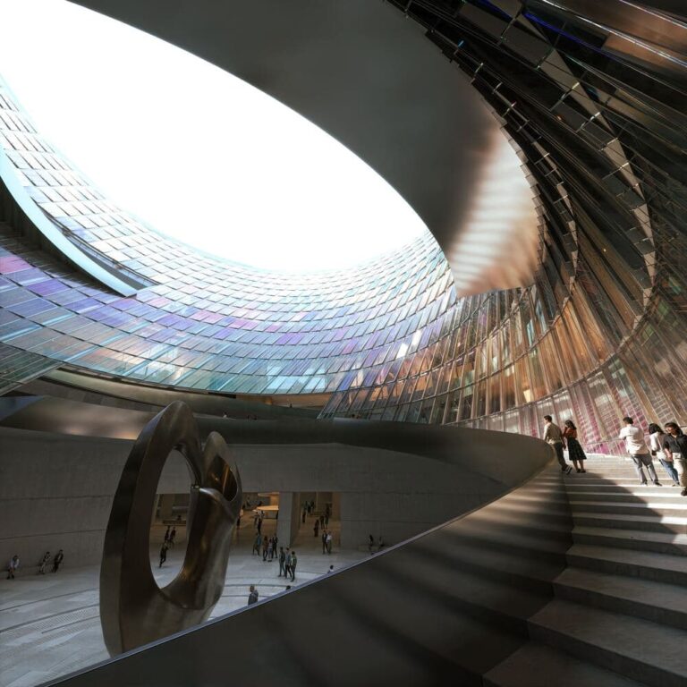 Interior view of the Beijing Art Museum’s atrium, featuring a sweeping curved staircase, a large sculptural installation, and a glazed dome that floods the space with natural light.
