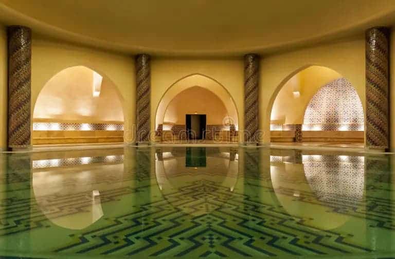 Modern reinterpretation of a Moroccan hammam interior with reflective pool, arched alcoves, and geometric zellige tilework  blending traditional Islamic spatial principles with contemporary wellness architecture for thermal and sensory comfort.
