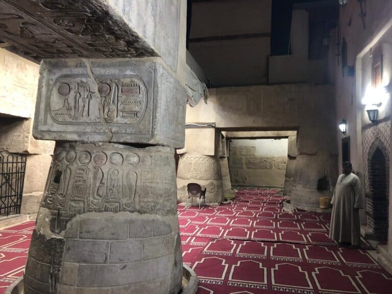 Interior of Abu Al-Haggag Mosque with carved Pharaonic columns featuring Ma’at symbols and prayer rugs, showing layered sacred architecture.