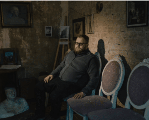 A bearded man with glasses and a grey zip-up jacket sits on a row of vintage oval-backed chairs in a dimly lit room with exposed brick walls, surrounded by sketches and sculptures.