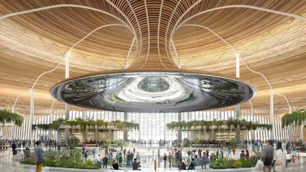 داخل محطة مطار جادوكدو الجديد، يبرز سقف خشبي نحتي وقرص عاكس مركزي، محاط بمساحات خضراء وحركة مسافرين.