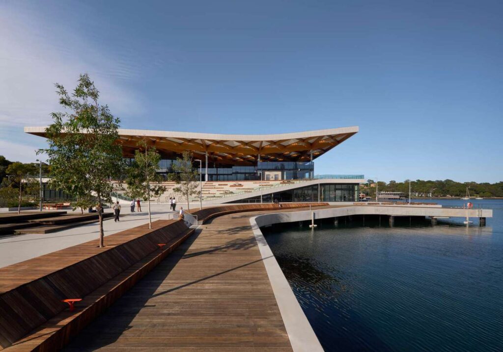 at Blackwattle Bay, viewed from a curved timber boardwalk along the waterfront, with its expansive roof and tiered terraces under clear blue sky.
