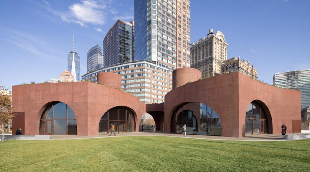 Wagner Park Pavilion in Lower Manhattan, featuring rust-colored concrete volumes with arched glass entrances and green roofs, set against a backdrop of skyscrapers including One World Trade Center &mdash; a fusion of flood resilience and civic architecture.