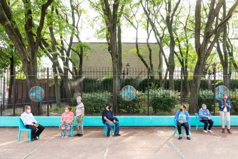 Community members seated on bright blue chairs in front of a black iron fence at Lopez Playground,   , with misting fans providing relief on a warm day.