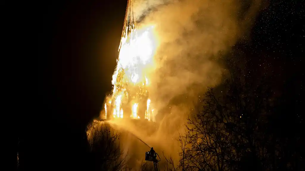 Firefighters battle a blaze engulfing the upper structure of a historic church tower at night, with water streams visible against the glowing flames. Architectural implications