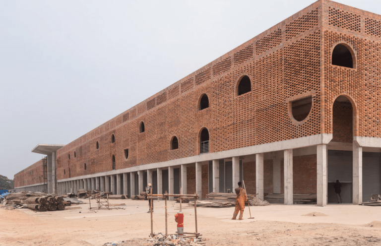 Perspective view of a large industrial building under construction featuring a massive red brick facade with intricate perforated patterns and arched openings atop a concrete pillar foundation.