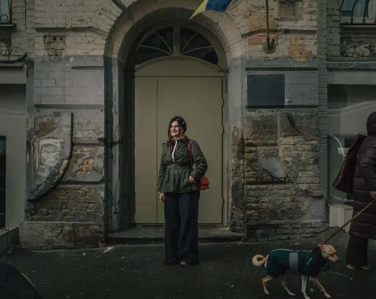 A woman in an olive green jacket and dark wide-leg trousers stands centered in front of a large, pale green door under a stone archway. A person walks a small dog in the foreground.