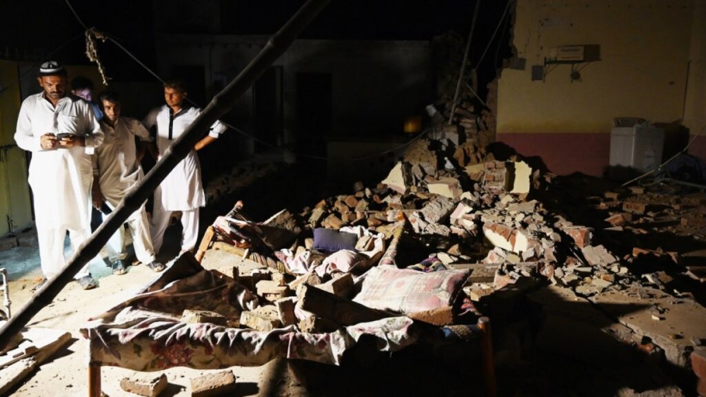 Survivors stand in the dark amidst the rubble of their destroyed home, illuminated by a single light source