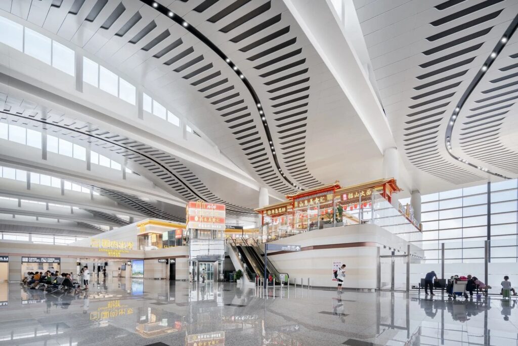 Interior showing curved ceiling with linear voids, domestic departures signage, and cultural food kiosks. Natural light floods the polished floor.