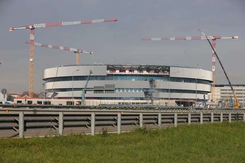 Wide-angle view of the Milan 2026 Olympic Ice Hockey Arena from across a highway, showing its curved metallic facade and active construction cranes under an overcast sky.