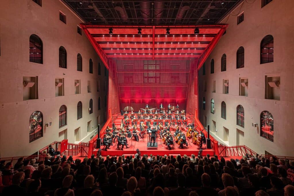 Interior of a concert hall with red-lit stage, orchestra performing, and audience seated in a repurposed industrial space with arched windows and exposed ceiling structure.