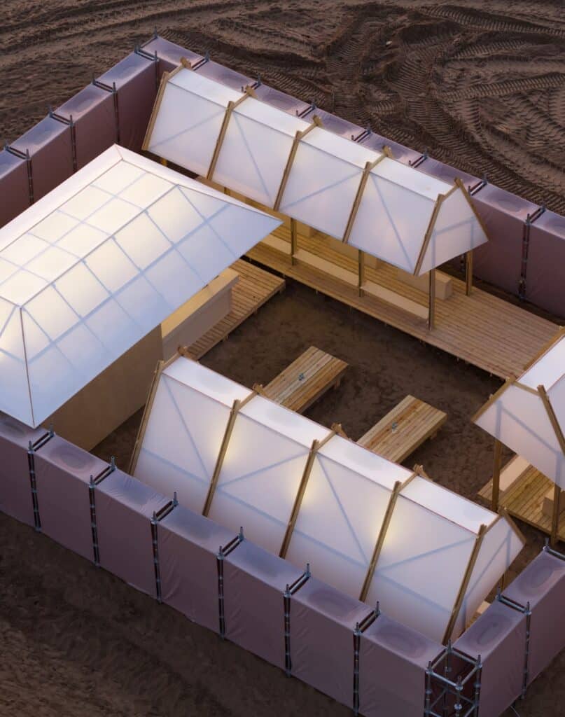 Overhead view of gabled roof modules and scaffolded perimeter walls forming a courtyard space on sandy ground.