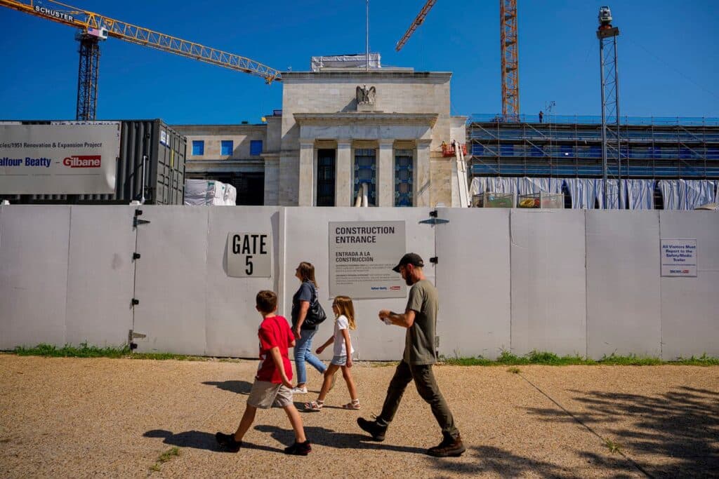 Construction fencing surrounds the Eccles Building in Washington, D.C., during its 2026 renovation, with cranes overhead and pedestrians walking past Gate 5 &mdash; illustrating urban integration of historic infrastructure upgrades.
