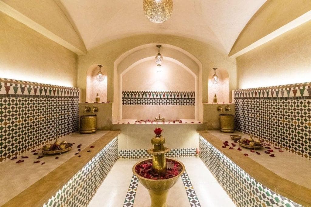 Luxury Moroccan hammam chamber with ornate zellige tilework, arched alcoves, and a central brass fountain adorned with rose petals &mdash; blending traditional Islamic design with modern wellness aesthetics for sensory relaxation.