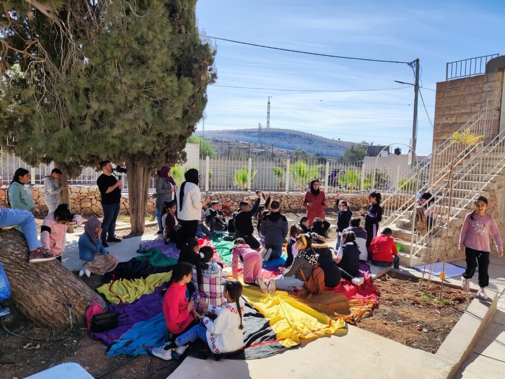 يجلس أطفال وبالغون تحت شجرة على بطانيات ملونة في الحديقة العامة بقلنديا، مع ظهور الجدار الفاصل في الخلفية.