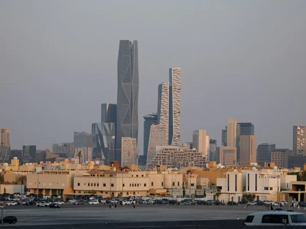 A wide shot of the King Abdullah Financial District (KAFD) skyline in Riyadh, featuring the PIF Tower and other geometric glass skyscrapers behind a row of traditional low-rise buildings and a parking area.