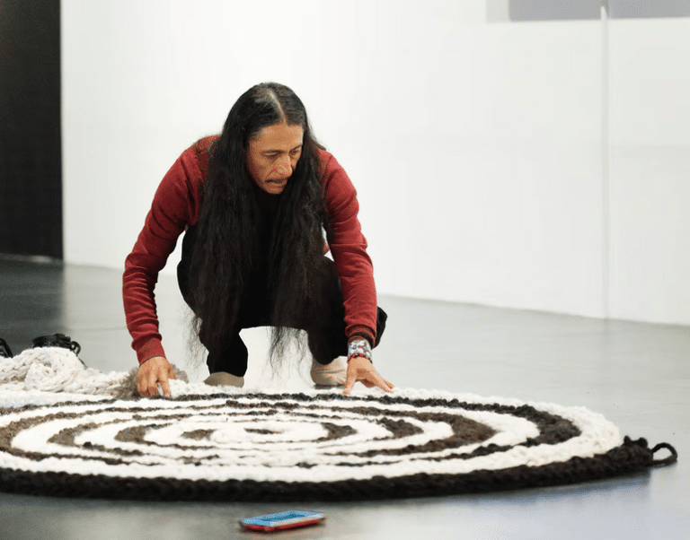 A man with long dark hair, wearing a red sweater, squats on a grey floor while meticulously arranging a large, circular, spiral-patterned rug made of thick black and white woven wool.
