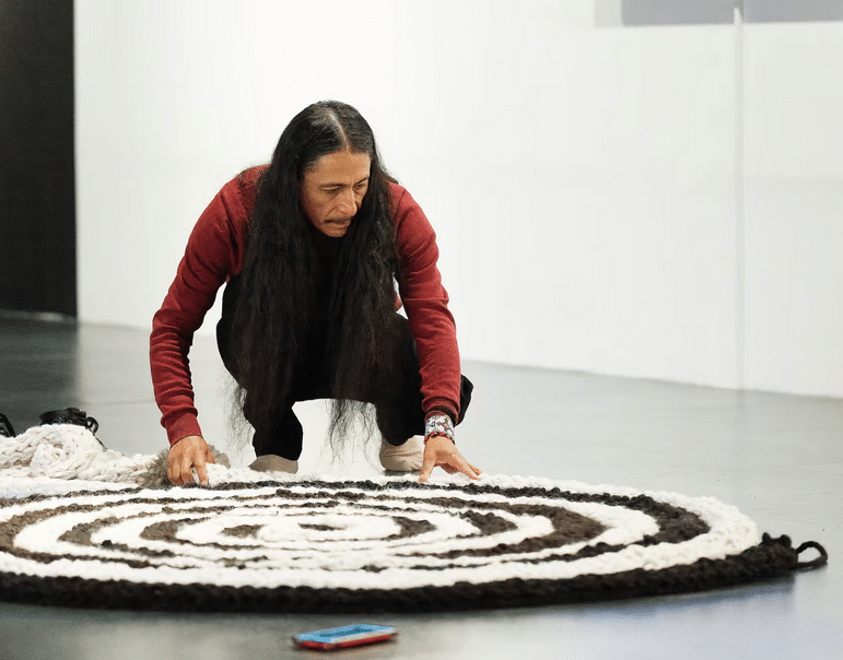 A man with long dark hair, wearing a red sweater, squats on a grey floor while meticulously arranging a large, circular, spiral-patterned rug made of thick black and white woven wool.
