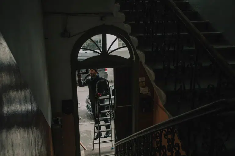A man standing on a silver stepladder in an arched doorway of an old building, framed by a dark stairwell with ornate wrought-iron railings.