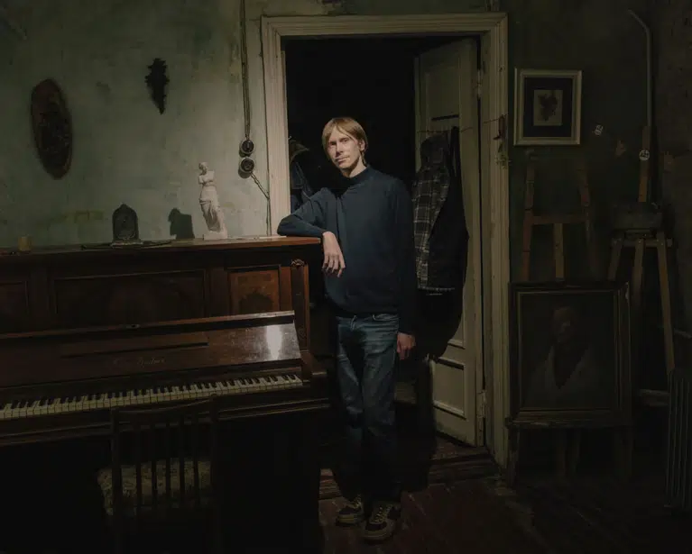 A tall, blonde man leaning on an upright piano in a dimly lit, vintage-style artist studio with teal walls, featuring a small white statue, an easel, and oil paintings.