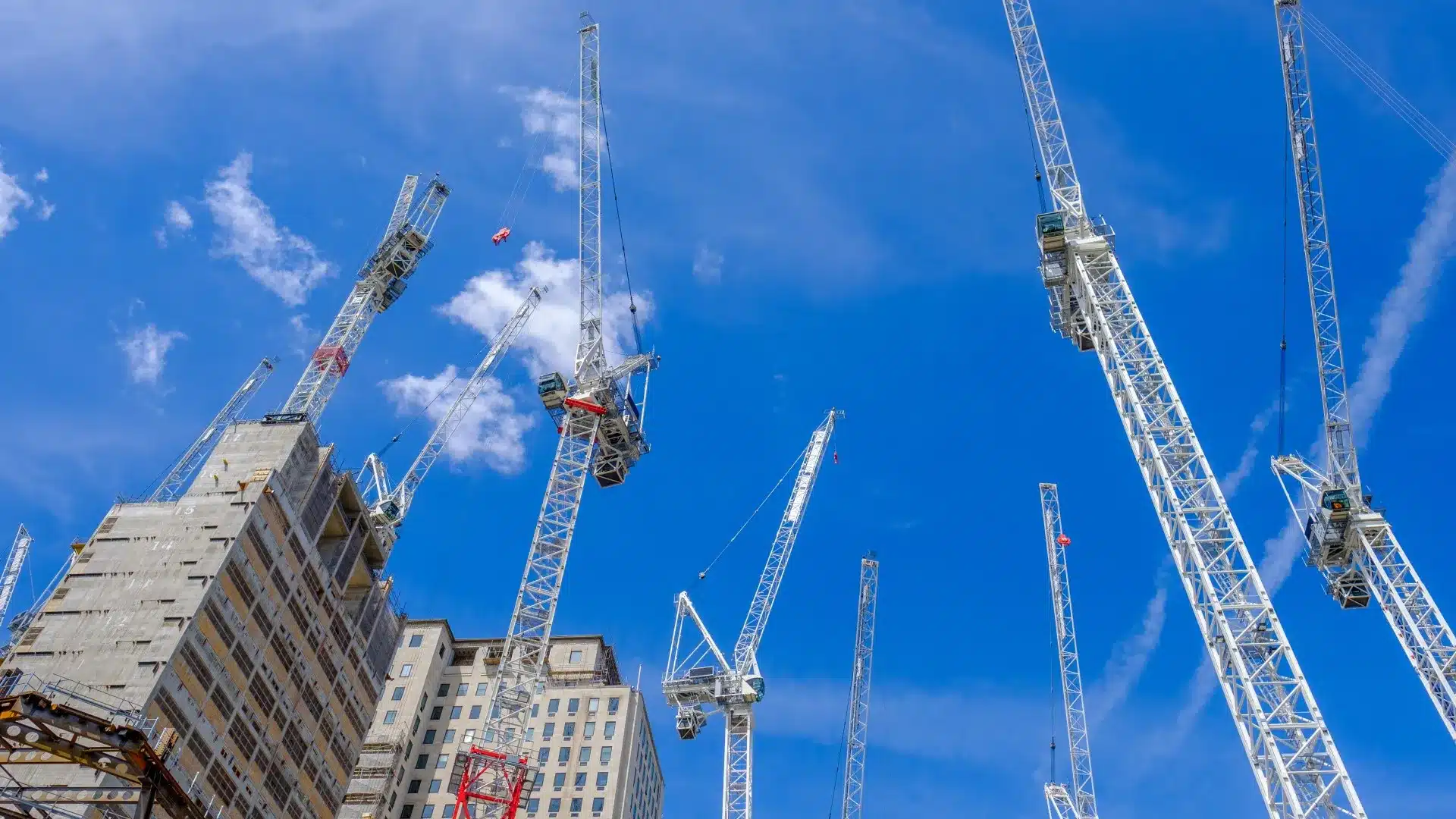 A low-angle view of multiple white luffing jib tower cranes working on a high-rise building construction site against a clear blue sky with light clouds.