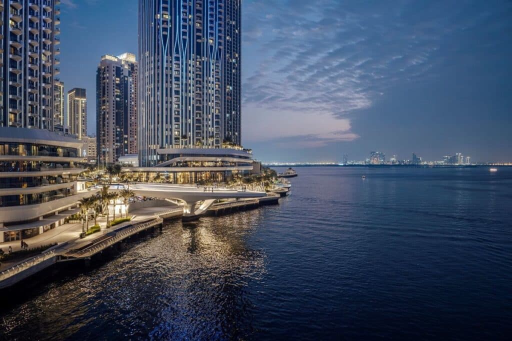 A high-angle evening view of Dubai Creek Harbour featuring illuminated luxury skyscrapers, a modern pedestrian promenade, and the calm water of the creek under a twilight sky.