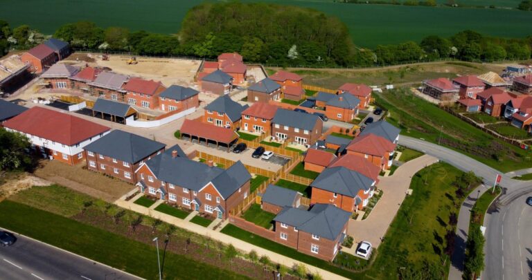 An aerial high-angle shot of a newly constructed residential neighborhood featuring brick houses with grey and red tiled roofs, surrounded by green fields and paved roads.