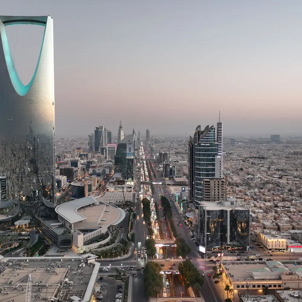 High-angle aerial view of King Fahd Road in Riyadh, featuring the Kingdom Centre skyscraper on the left and heavy traffic flowing through the city at sunset.